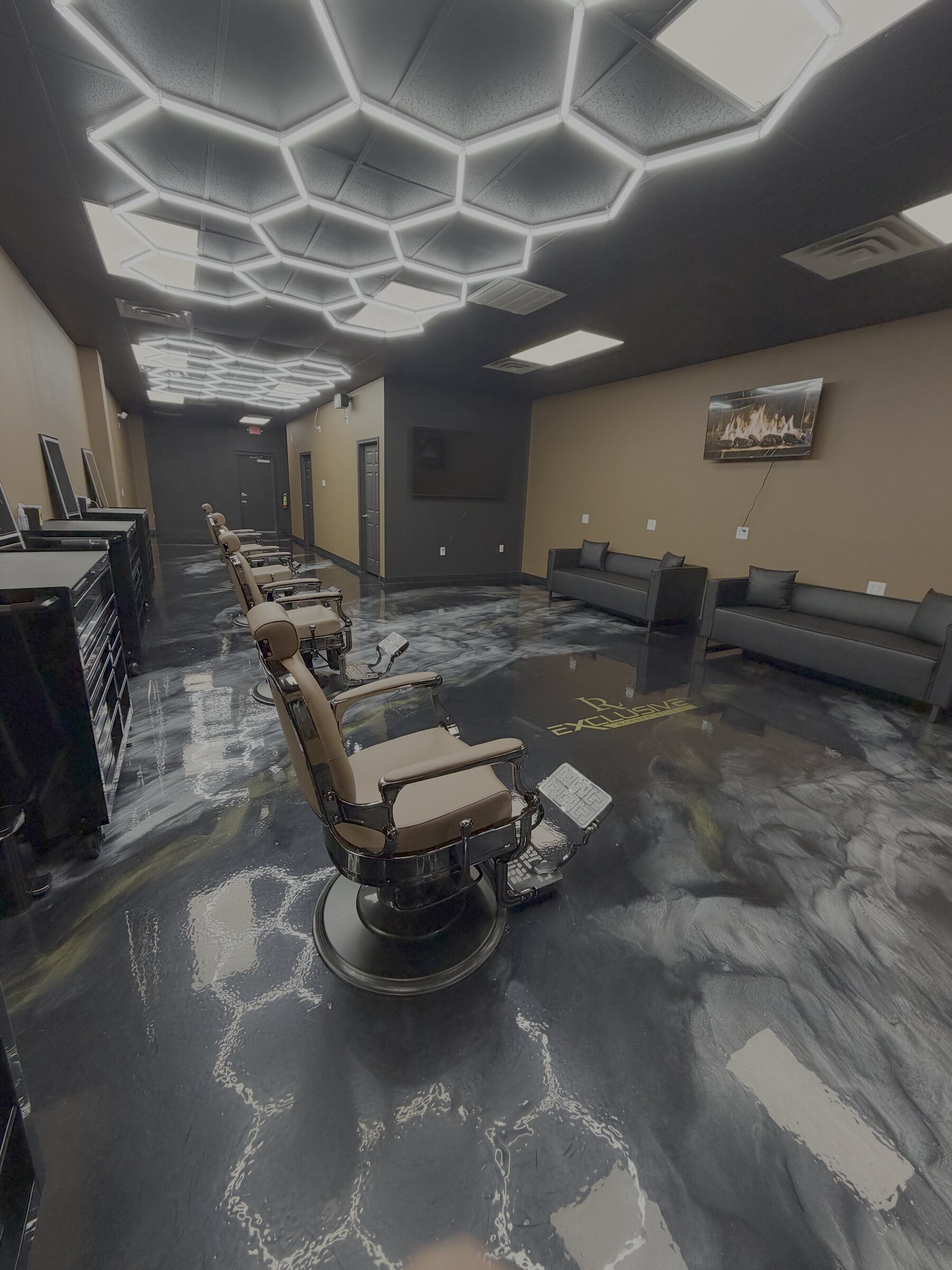 Exclusive Barber Studio interior — hexagonal LED ceiling, marbled epoxy floor with the gold studio mark, vintage barber chairs and a black leather lounge.
